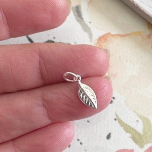 May include: A small silver leaf charm with a detailed vein pattern. The charm is shown held in a hand.