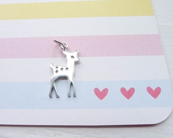 Peut inclure: Un charme en argent en forme de cerf tacheté. Le charme est sur un fond blanc avec des rayures roses et bleues.