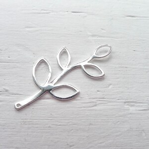 May include: Silver metal leaf branch charm with a delicate design. The branch has five leaves with cut-out details. The charm has a loop at the end for attaching to jewelry or other crafts.