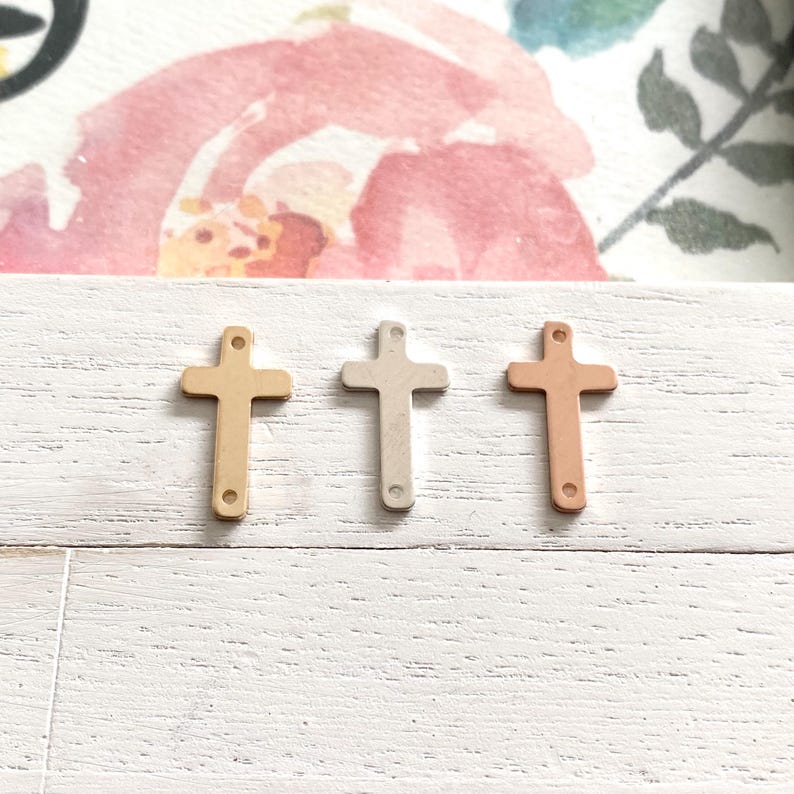 May include: Three small metal crosses in different finishes: gold, silver, and rose gold. The crosses are arranged in a row on a white wooden surface.