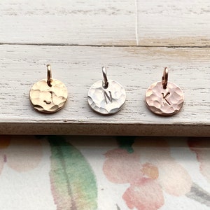 May include: Three hammered metal disc pendants with the letters J, N, and K. The pendants are gold, silver, and rose gold, each with a small loop for hanging. The background is a white wooden surface.