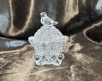 May include: A clear glass candy dish with a lid featuring a bird finial. The bowl and lid are decorated with a pressed diamond and starburst pattern. The base of the dish is supported by three small, curved feet, creating a stable foundation.