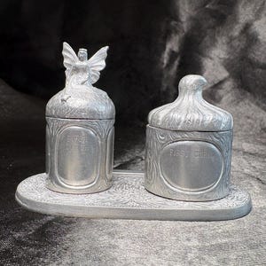 May include: A silver-colored keepsake set featuring a fairy-topped box labeled "FIRST TOOTH" and a bird-topped box labeled "FIRST CURL". The set sits on an oval tray with an embossed floral design. A charming gift.