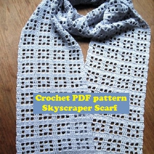 May include: A light blue crochet scarf with a lacy pattern. The scarf is folded in half and laid on a wooden surface. The text "Crochet PDF pattern Skyscraper Scarf" is printed on a blue and yellow banner.