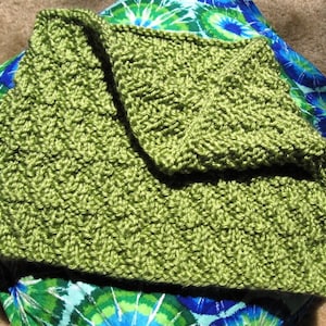 May include: A green knitted blanket folded in half, with a textured pattern. The blanket is lying on a blue and green patterned fabric.