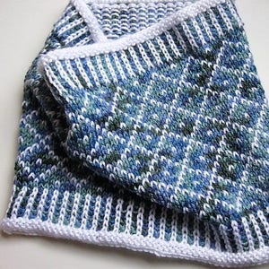 Peut inclure: Une écharpe en tricot blanche et bleue avec un motif géométrique. L'écharpe est pliée en deux et a une bordure blanche.