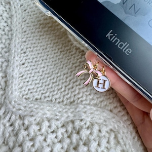 May include: A gold charm with a pink bow and a white circle with the letter "H" in gold. The charm is attached to a black Kindle e-reader.