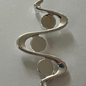 May include: Silver pendant with three round cutouts. The pendant is shaped like a stylized letter S.
