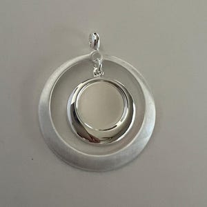 May include: A silver pendant featuring two nested circular shapes. The outer ring has a brushed finish, while the inner ring is highly polished, creating a contrast in texture and shine. The pendant hangs from a small loop.