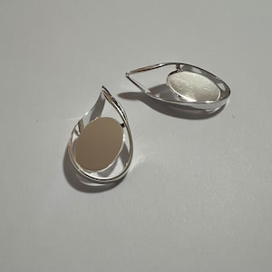 May include: Two silver-toned teardrop-shaped pendants. Each pendant features a polished, oval-shaped center within a curved frame. The pendants are displayed on a light gray surface, showcasing their reflective qualities and minimalist design.