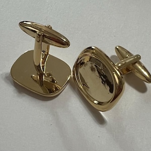 May include: A pair of gold cufflinks. One cufflink is a square shape with a smooth surface. The other cufflink is a rounded oval shape with a textured surface.
