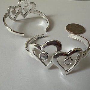 May include: Two silver-colored heart-shaped cuff bracelets. Each bracelet features two connected hearts with a small, round setting in the center of each heart. A coin is next to one of the bracelets.