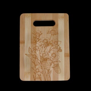 May include: A natural bamboo cutting board with a floral design engraved on the surface. The design features a bouquet of flowers and strawberries.