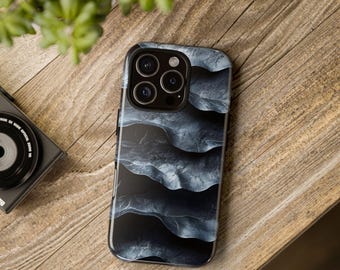 May include: A smartphone case with a dark, abstract design featuring layered, wave-like patterns in shades of gray and black. The case is on a wooden surface, with a camera and plant in the background.