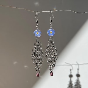 Puede incluir: Pendientes colgantes con flores azules de nomeolvides encapsuladas en resina transparente. Los pendientes tienen un diseño de cota de malla plateada y están acabados con pequeñas cuentas rosas.
