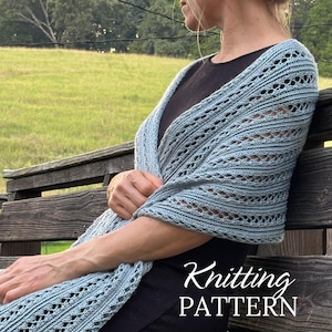 May include: A light blue knitted shawl with a delicate openwork pattern is draped over a person's shoulders. The text "Knitting PATTERN" is visible in a cursive font. The shawl is worn over a black top.