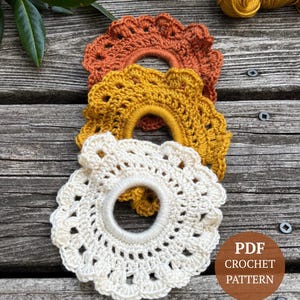 May include: Three handmade crochet hair scrunchies in cream, mustard yellow, and burnt orange. The scrunchies are circular with a scalloped edge. The image also includes a brown circle with the text "PDF CROCHET PATTERN".