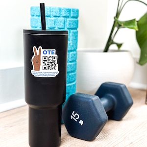 May include: A black tumbler with a straw and a sticker that says "VOTE" with a QR code. A blue foam roller and a 15-pound dumbbell are also in the image. The items are on a light wood surface.