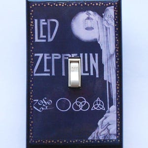 May include: A decorative light switch cover featuring the band name "LED ZEPPELIN" in white lettering on a dark blue background. The cover includes a white switch and a graphic of a figure holding a lantern. The border is black with gold stars.