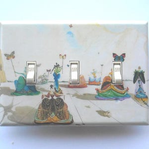 May include: A decorative light switch cover featuring a surrealist painting with butterflies and figures. The cover has three silver toggle switches, each labeled "OFF". The artwork includes a variety of colors, including blue, green, and orange.