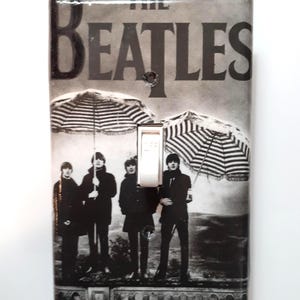 May include: A light switch cover featuring a black and white image of The Beatles. The image shows the band members standing under striped umbrellas. The word "BEATLES" is printed at the top.