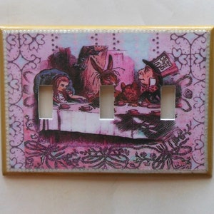 May include: A decorative light switch cover featuring a colorful illustration of the Mad Hatter's tea party from Alice in Wonderland. The cover has three switch openings and a gold border. The background is pink with decorative floral elements.