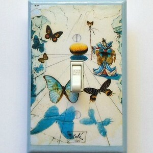 May include: Light blue wall switch plate featuring a surreal butterfly design with a white toggle switch. The artwork includes butterflies in shades of blue and brown, with a light blue border. The word "Dali" is visible in the lower part of the plate.
