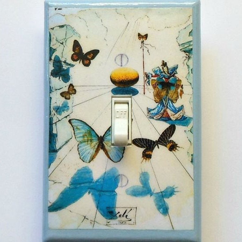 May include: Light blue wall switch plate featuring a surreal butterfly design with a white toggle switch. The artwork includes butterflies in shades of blue and brown, with a light blue border. The word "Dali" is visible in the lower part of the plate.