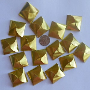 May include: A collection of gold-colored, square-shaped decorative elements. Each square has a folded design, with four curved sections meeting in the center. A quarter coin is included for size reference. The elements are arranged on a white surface.
