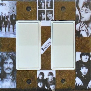 May include: A decorative light switch cover featuring images of The Beatles. The cover has two light switch openings with a brown background. The Beatles' band name is printed on a white label. Photos of the band members are arranged around the switches.