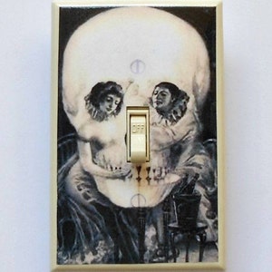 May include: A decorative light switch cover featuring a skull design with two figures. The artwork is in black and white, with the word "OFF" visible on the switch. The cover has a cream-colored border.