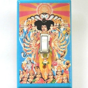 May include: A light blue wall switch cover featuring a colorful illustration. The artwork depicts a figure with multiple arms and a face resembling Jimi Hendrix, surrounded by other figures. The switch is in the "off" position.