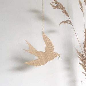 Peut inclure: Une décoration d'oiseau en bois en forme d'hirondelle, suspendue à une chaîne dorée. L'oiseau est brun clair et a une petite perle verte suspendue à son bec.