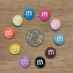 May include: A collection of colorful, round candies with the letter "m" printed on them, arranged around a quarter coin. The candies are in various colors, including blue, pink, orange, yellow, and purple.