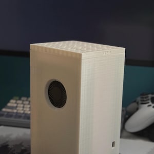 Puede incluir: Una caja blanca impresa en 3D con un ventilador negro y un pequeño puerto en el lateral. La caja tiene una superficie texturizada y una pequeña etiqueta en la parte superior que dice "LUMI".