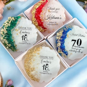 May include: Four small white boxes with gold accents, each containing a round, clear resin favor with a different color and design. The favors have text on them, including names, ages, and dates. The favors are for a birthday party or other special event.