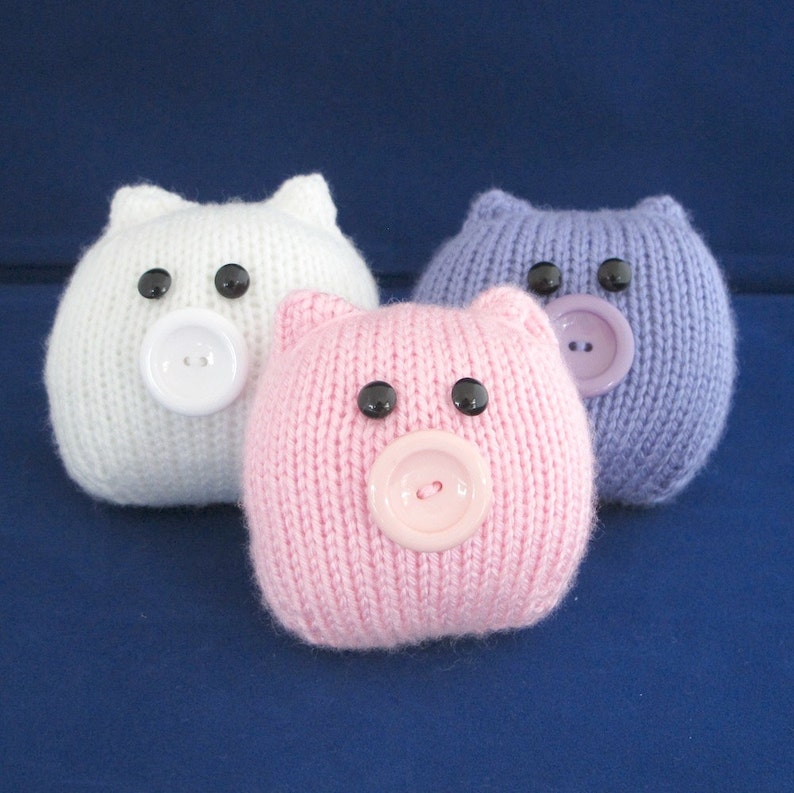 May include: Three knitted pig toys, one white, one pink and one purple. Each pig has black eyes and a button nose.