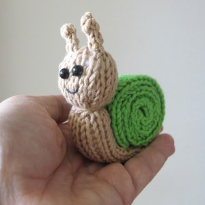 May include: A knitted snail toy with a beige body and green shell. The snail has black eyes and a small smile.