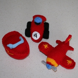 Könnte beinhalten: Drei gestrickte Spielzeuge: ein rotes Rennauto mit schwarzen Rädern und der Nummer 4, ein rotes Flugzeug mit gelben Akzenten und einem blauen Propeller und ein rotes Boot mit einem blauen Griff.