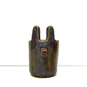Könnte beinhalten: Eine kleine, handgefertigte Keramikvase mit einer einzigartigen Form. Die Vase ist tiefblau mit schillernden Gold- und Lilatönen. Sie hat zwei Griffe oben und ein kleines rechteckiges Detail auf der Vorderseite.