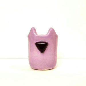 May include: A lavender ceramic cat-shaped vase with a dark brown triangular accent. The vase has two pointed ears at the top and a smooth, glossy finish. The cat-themed design is simple and modern.