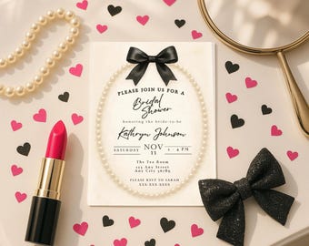 May include: A bridal shower invitation with a black bow, surrounded by a pearl necklace. A tube of red lipstick and a black glitter bow are also present. Pink and black heart confetti decorates the white surface.
