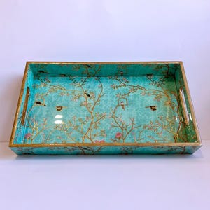 May include: Rectangular turquoise serving tray with gold trim and handles. The tray features a detailed design of birds, flowering branches, and birdhouses. The tray's interior has a textured pattern.