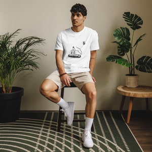 Könnte beinhalten: Weißes T-Shirt mit einer schwarzen Strichzeichnung einer meditierenden Figur und Rauch. Das Shirt hat die Buchstaben "KB" in Schwarz. Die Person trägt beige Shorts und weiße Turnschuhe. Die Person sitzt auf einem Hocker.