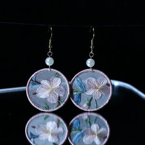 Può includere: Orecchini floreali ricamati. Ogni orecchino presenta un design rotondo in tessuto grigio con un fiore rosa e bianco e foglie blu. I bordi sono rifiniti con filo rosa. Una piccola perla e un gancio dorato completano il design.