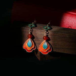 May include: A pair of ornate earrings with a layered, fan-shaped design in orange, red, and turquoise. Each earring features a red bead and a green decorative top, suspended from a gold-colored hook.