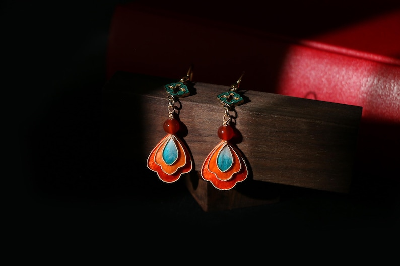 May include: A pair of ornate earrings with a layered, fan-shaped design in orange, red, and turquoise. Each earring features a red bead and a green decorative top, suspended from a gold-colored hook.