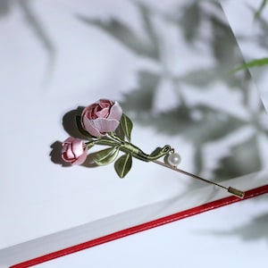 May include: A delicate floral brooch featuring two pink rose blossoms with green leaves and a pearl accent. The brooch is attached to a long, gold-toned pin. The design is set against a white background.