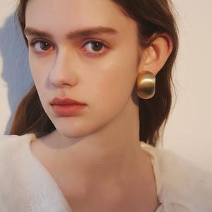May include: A thick, curved gold-toned hoop earring with a smooth, matte finish. The earring has a wide, rounded profile that sits flush against the earlobe, featuring a solid metallic construction with a soft, reflective surface texture.