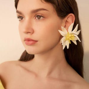 May include: A large statement earring featuring a white flower with pointed petals and a cluster of yellow beads at the center. The earring is worn on the earlobe, showcasing a detailed floral design with contrasting white and yellow colors.
